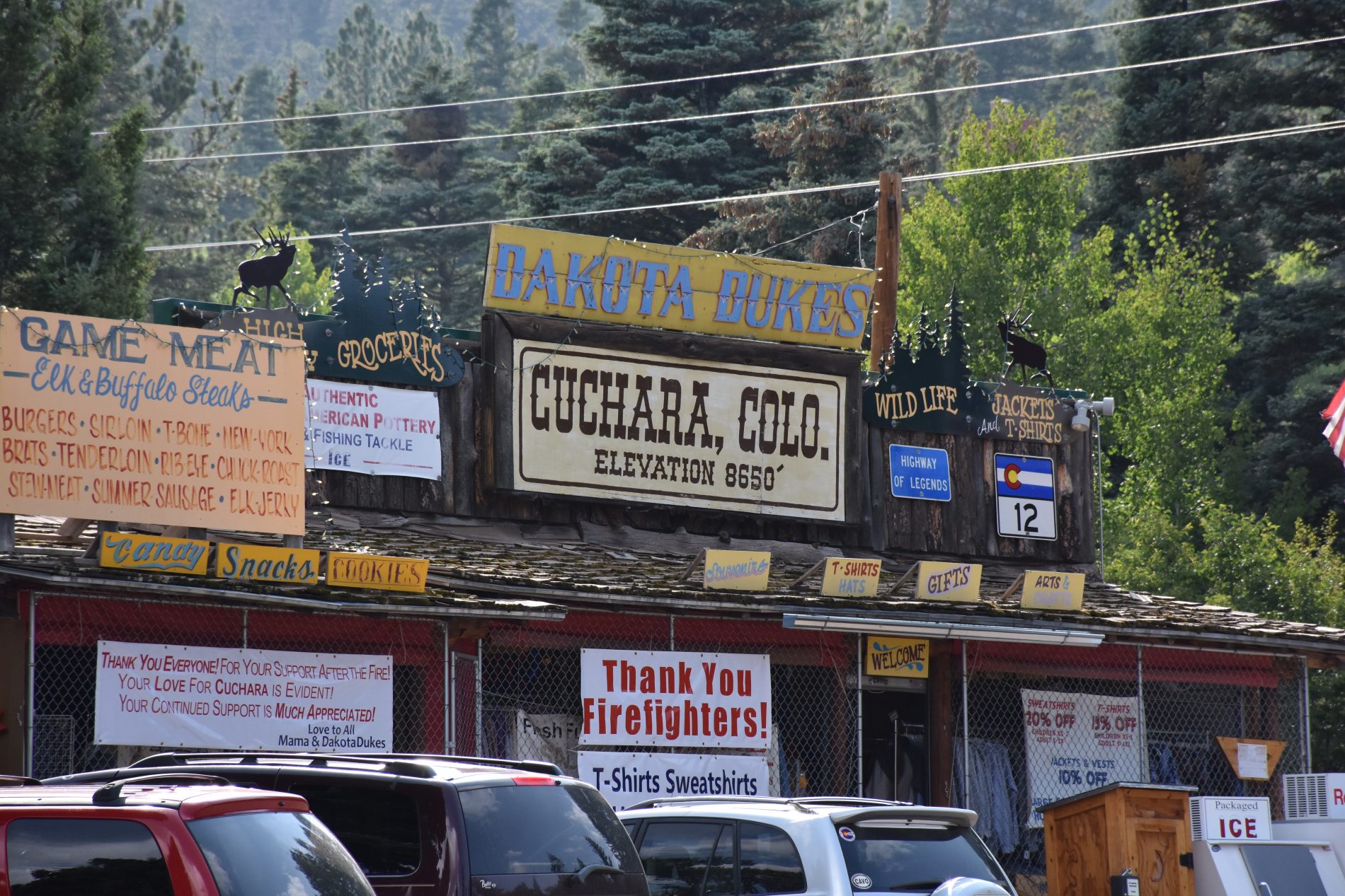 A log built building, racks of shirts show, a white banner with red printing reads thank you firefighters!. Yellow sign with blue lettering reads Dakota Dukes. Brown writing below reads Cuchara, Colorado. Elevation 8650.