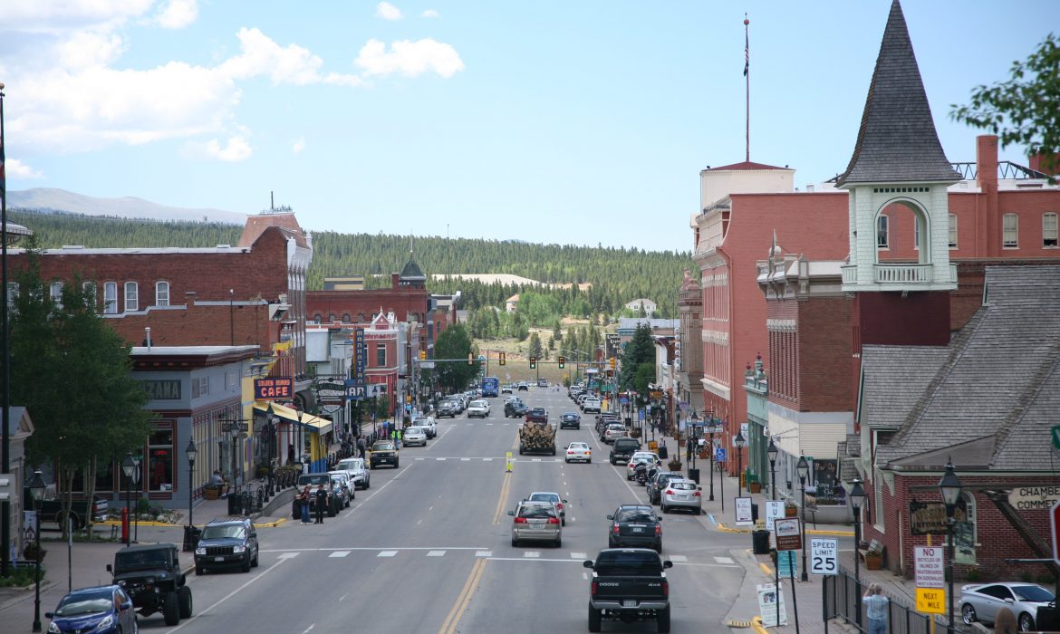 A busy downtown street lined with brick two and three story buildings. Cars parked on both sides of street, people walking on sidewalk. View extends to pine tree studded hills, and high mountain peaks.